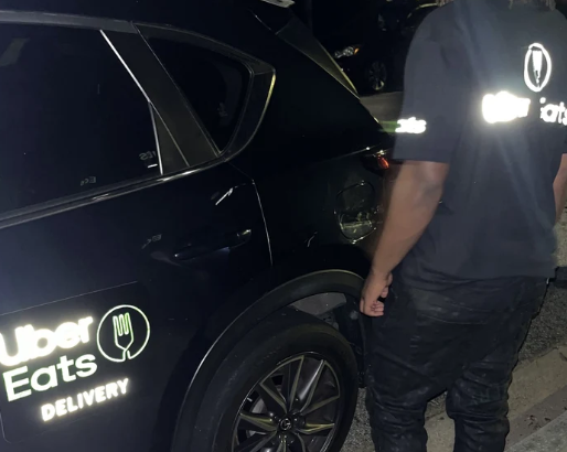 Person wearing a reflective Uber Eats Tshirt standing next to a black car with a Reflective Uber Eats at night.